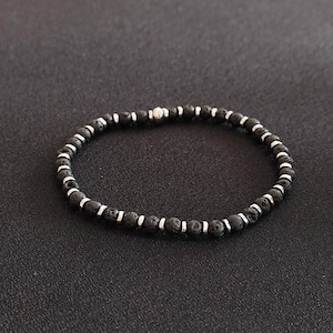 Könnte beinhalten: Schwarzes Perlenarmband mit silberfarbenen Akzenten. Das Armband besteht aus kleinen, runden, schwarzen Perlen, die mit silberfarbenen Abstandshaltern durchsetzt sind. Eine einzelne, größere silberne Perle ist zentriert. Das Armband wird auf dunklem Untergrund präsentiert.