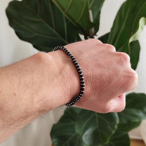 Könnte beinhalten: Schwarzes Perlenarmband mit silberfarbenen Akzenten. Das Armband besteht aus kleinen, runden schwarzen Perlen, die durch dünne, silberfarbene Abstandshalter getrennt sind. Das Armband wird am Handgelenk getragen.