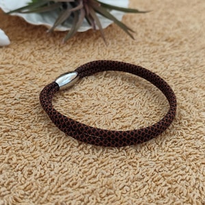 Könnte beinhalten: Ein braun-schwarz gemustertes Armband mit einem silbernen Magnetverschluss. Das Armband ist kreisförmig und scheint aus gewebtem Stoff zu bestehen. Der Verschluss hat eine glänzende, metallisch silberne Farbe.