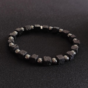 Könnte beinhalten: Ein schwarzes Armband mit quadratischen, strukturierten Lavasteinperlen, die mit glatten, silberfarbenen Perlen abwechseln. Das Armband wird vor einem dunklen Hintergrund gezeigt, um den Kontrast hervorzuheben.