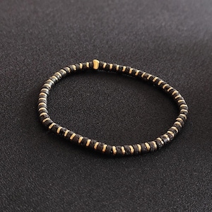 Könnte beinhalten: Schwarzes Perlenarmband mit goldfarbenen Akzenten. Das Armband besteht aus kleinen, runden schwarzen Perlen, die mit goldfarbenen zylindrischen Perlen durchsetzt sind. Das Armband wird auf einer dunklen Oberfläche präsentiert.