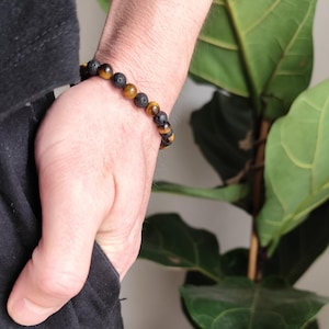 Könnte beinhalten: Ein Armband mit schwarzen Lavastein- und Tigeraugen-Perlen. Das Armband wird am Handgelenk getragen und zeigt die natürlichen Farben und Texturen der Steine. Das Armband ist ein Schmuckstück.