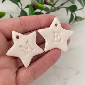 May include: Two white ceramic star ornaments with the letters "M" and "E" embossed on them. The stars are held in a hand.