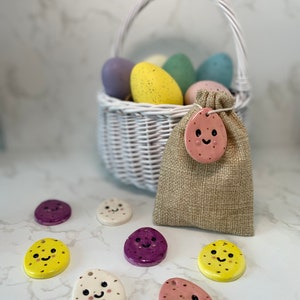 May include: A white wicker basket filled with pastel coloured speckled Easter eggs. A small burlap bag with a pink ceramic egg ornament with a happy face is next to the basket.  There are seven ceramic egg ornaments with happy faces in various colours on a white marble surface.