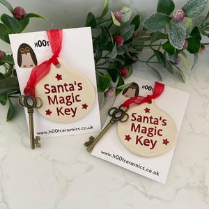 May include: Two white ceramic ornaments shaped like keys with red text that reads "Santa's Magic Key" and a red star on each side of the text. The ornaments are hanging from red ribbon and are on white cards with the website "www.hootceramics.co.uk" printed on them.
