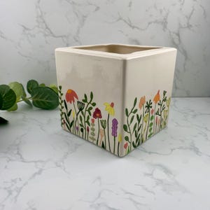 May include: A square, cream-coloured ceramic planter with a colourful floral design. The planter features a border of stylised flowers in shades of red, yellow, orange, and purple, with green stems and leaves. Suitable for indoor or outdoor use.