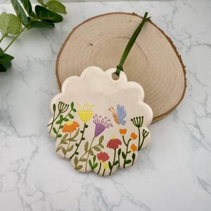 May include: A cream-coloured, scalloped-edge ceramic ornament with a colourful floral design and a butterfly. The flowers are in shades of orange, yellow, purple, and red. A green ribbon is attached for hanging.