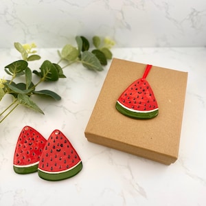 May include: Three ceramic watermelon slice ornaments. Two are small and have a smiling face. One is larger and hangs from a red string. All are red with black seeds and green rind.
