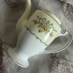 Peut inclure: Théière en porcelaine vintage avec un corps crème et un motif floral. La théière présente un bec verseur incurvé, une anse et une bordure dorée. Le motif floral comprend des fleurs roses et bleues avec des feuilles vertes.