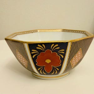 May include: An octagonal ceramic bowl with a white interior and a navy blue exterior featuring a red floral design. The bowl has gold trim and a lattice pattern, adding a touch of elegance to its design.