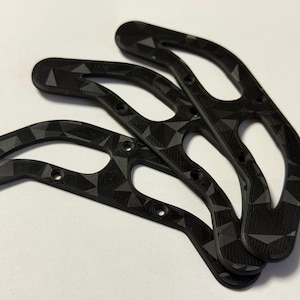 Könnte beinhalten: Drei schwarze, geometrisch gemusterte, gebogene Halterungen mit ovalen Öffnungen und kleinen kreisförmigen Löchern. Die Halterungen sind leicht überlappend angeordnet und zeigen ihr kompliziertes Design und ihre Textur. Der Hintergrund ist eine schlichte weiße Oberfläche.