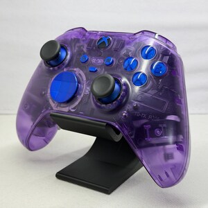 Könnte beinhalten: Ein transparent-violetter Videospiel-Controller mit blauen Akzenten und Knöpfen, der auf einem schwarzen Ständer steht. Der Controller hat ein klares Design, das die internen Komponenten zeigt. Das Xbox-Logo ist auf dem Controller sichtbar.