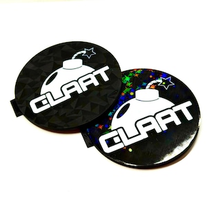 May include: Two black round stickers with the word "CLAAT" in white, and a bomb graphic. One sticker has a geometric pattern, the other has a holographic effect with colorful stars.