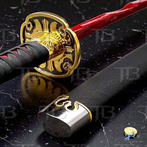 May include: A katana sword is shown in detail. The sword has a red handle and a gold-coloured guard with detailed patterns. The handle is wrapped in black material, and the scabbard is black. The blade is not visible.