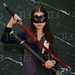 Könnte beinhalten: Ein schwarzes und rotes Katana-Schwert mit einem schwarzen Griff und einer roten Klinge. Das Schwert befindet sich in einer schwarzen Scheide mit goldenen Akzenten. Die Person trägt eine schwarze Maske.