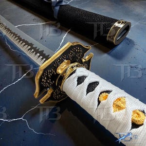 May include: Close-up of a katana sword. The silver blade has a serrated edge. The hilt is wrapped in white fabric with black and gold details. The black scabbard features gold accents. A traditional Japanese weapon.