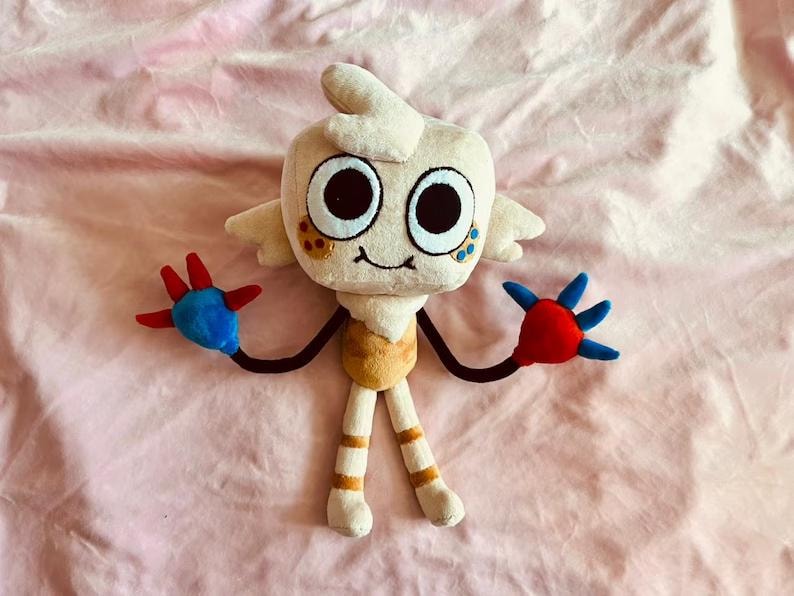 May include: A plush toy character with a cream-colored head, large black eyes, and small wings. The toy has black arms ending in red and blue hands. The body is tan and white, with tan and orange striped legs.