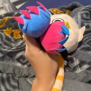 May include: A plush toy featuring a blue and a pink orb with red and blue accents, held in a hand. The toy has a white face with yellow details and a striped yellow and white tail. The background is a patterned blanket.