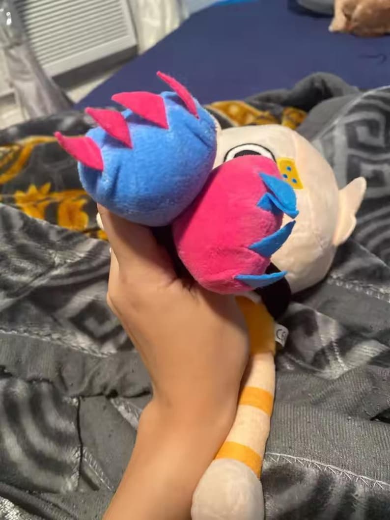 May include: A plush toy featuring a blue and a pink orb with red and blue accents, held in a hand. The toy has a white face with yellow details and a striped yellow and white tail. The background is a patterned blanket.