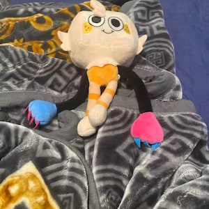May include: A plush toy with a cream-colored head, orange striped legs, and black arms. The toy has a smiling face with large eyes and small yellow accents. The arms end in blue and pink claw-like shapes. The toy is resting on a gray blanket.