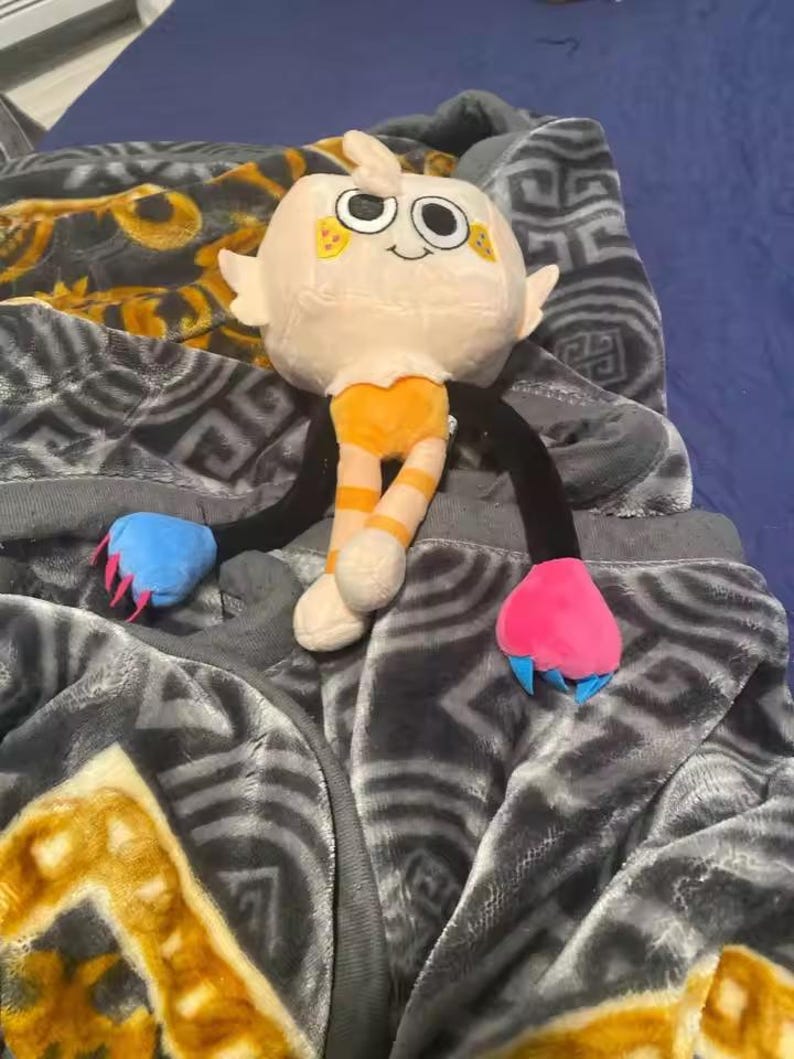 May include: A plush toy with a cream-colored head, orange striped legs, and black arms. The toy has a smiling face with large eyes and small yellow accents. The arms end in blue and pink claw-like shapes. The toy is resting on a gray blanket.