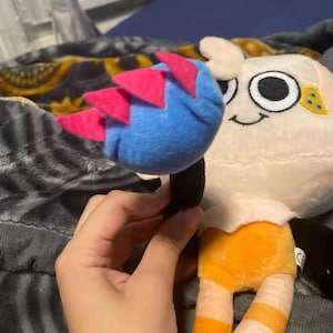 May include: A plush toy featuring a character with a cream-colored head, large black eyes, and a smiling mouth. The character has a blue and pink headpiece and orange and white striped legs. The toy is held by a hand.