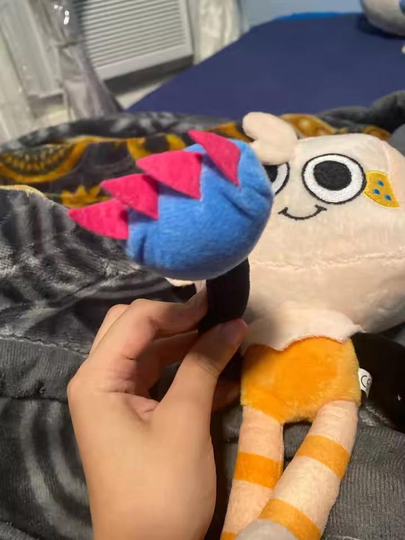May include: A plush toy featuring a character with a cream-colored head, large black eyes, and a smiling mouth. The character has a blue and pink headpiece and orange and white striped legs. The toy is held by a hand.