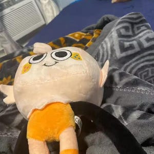 May include: A plush toy with a round, off-white head featuring large, black eyes and a small smile. The toy has orange shorts and striped legs, with long black arms. The toy is lying on a patterned blanket.