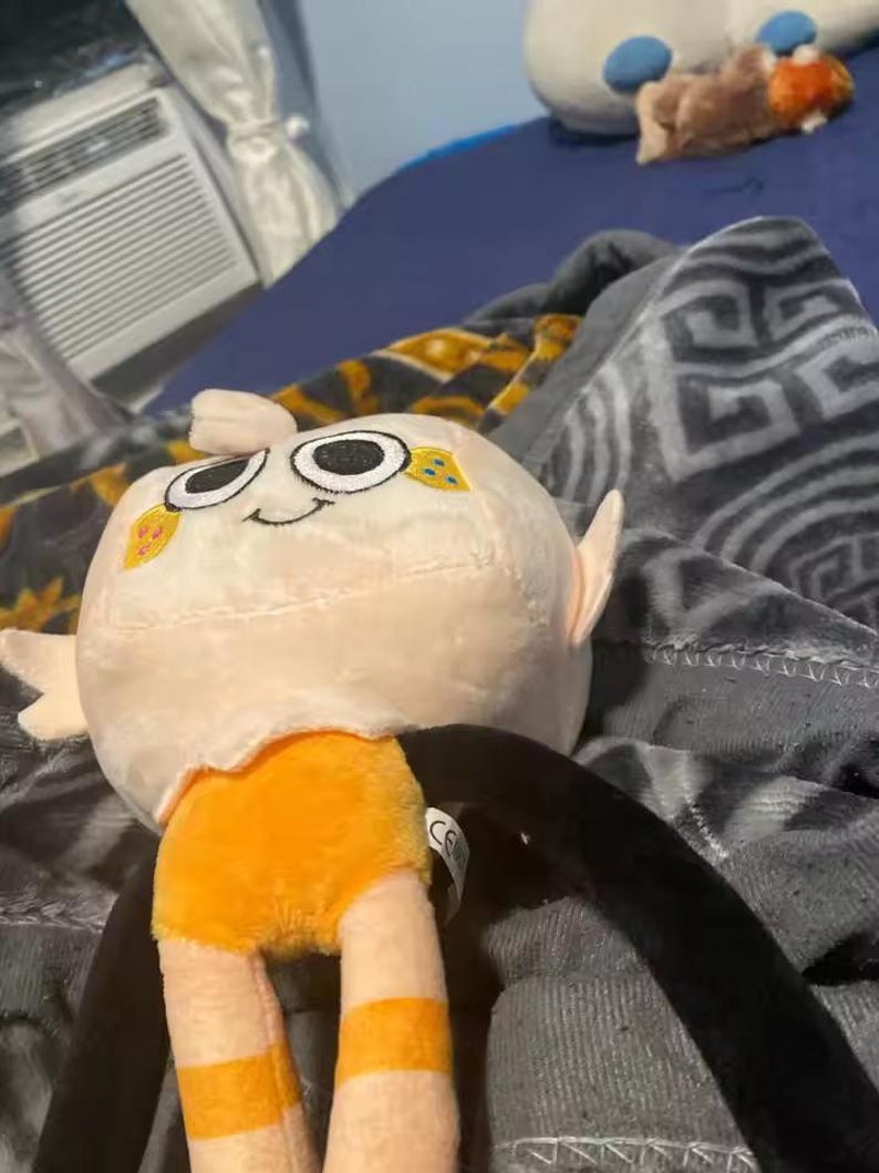 May include: A plush toy with a round, off-white head featuring large, black eyes and a small smile. The toy has orange shorts and striped legs, with long black arms. The toy is lying on a patterned blanket.