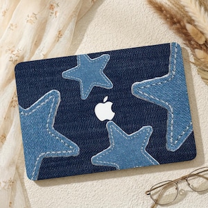 May include: A navy blue laptop case featuring four light blue denim stars with white stitching. The Apple logo is visible in the center. The case is on a light beige surface with glasses and decorative elements.