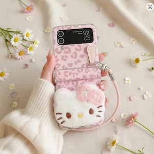 May include: Pink leopard print phone case with a fluffy Hello Kitty design. The case has a wrist strap and a clear protective cover. Daisies and flower petals are scattered around the case.