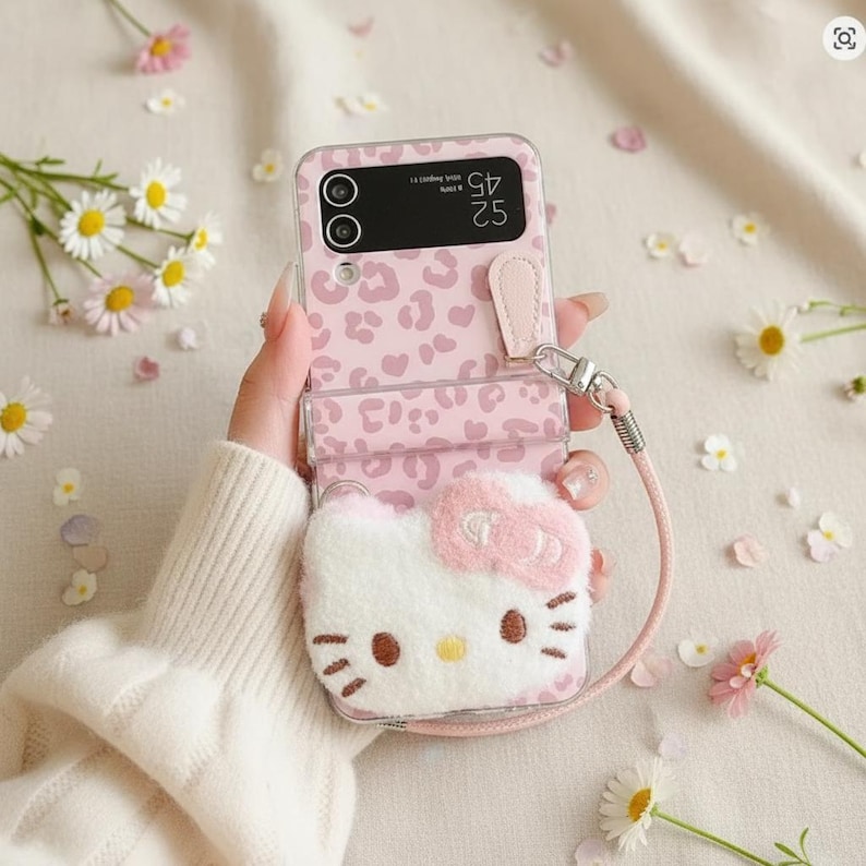 May include: Pink leopard print phone case with a fluffy Hello Kitty design. The case has a wrist strap and a clear protective cover. Daisies and flower petals are scattered around the case.