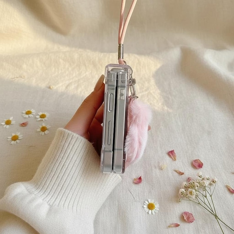 May include: Clear phone case with a pink lanyard and a fluffy pink keychain. The phone case is held vertically, showcasing its transparent design. The background features scattered flowers and petals.