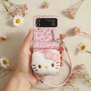 May include: Pink phone case with a leopard print design and a white Hello Kitty plushie attached. The case has a pink strap and is held in a hand, surrounded by flowers.