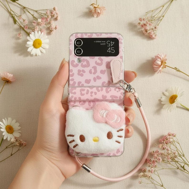 May include: Pink phone case with a leopard print design and a white Hello Kitty plushie attached. The case has a pink strap and is held in a hand, surrounded by flowers.