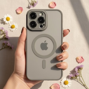 Puede incluir: Una funda gris para teléfono con un diseño MagSafe circular y un logotipo de Apple. La funda tiene un recorte para varias lentes. La funda del teléfono está sujeta por una mano, rodeada de flores y pétalos.