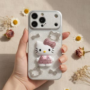 Puede incluir: Funda de tel&eacute;fono plateada con un dise&ntilde;o 3D de Hello Kitty, con un lazo y un vestido rosas, y adornos de pedrer&iacute;a. La funda tiene un fondo brillante y detalles adicionales de lazo y Hello Kitty de pedrer&iacute;a.