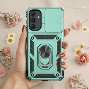 Puede incluir: Funda de teléfono verde menta con tapa de cámara y soporte de anillo. La funda tiene un diseño geométrico con detalles en negro y un agarre texturizado. El soporte del anillo es plateado y circular.