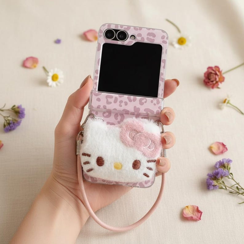 May include: A pink and clear phone case with a leopard print design, featuring a plush Hello Kitty character. The case has a strap and is open, revealing the phone's screen. Floral accents surround the phone.