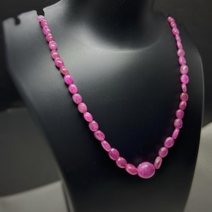 Puede incluir: Un collar de piedras preciosas rosas expuesto sobre un maniquí negro. El collar presenta una serie de piedras preciosas rosas pulidas de forma ovalada, con una piedra preciosa más grande y redonda en el centro. Las piedras están ensartadas, creando una joya elegante.