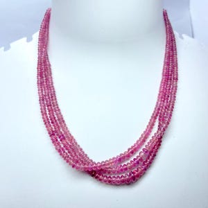 May include: A multi-strand necklace composed of small, round, pink gemstones. The necklace is draped on a white mannequin bust, showcasing the delicate beads and the overall design. The pink color is consistent throughout the necklace.