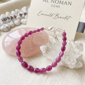 Peut inclure: Un bracelet composé de petites perles ovales de couleur magenta profond enfilées sur une chaîne argentée avec un fermoir mousqueton. Une carte de présentation derrière le bracelet porte l'inscription AL NOMAN GEMS et Emerald Bracelet.