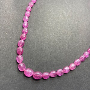 Puede incluir: Un collar compuesto por piedras preciosas rosas ovaladas y pulidas. Las cuentas están ensartadas, creando un bucle continuo. Las piedras preciosas son translúcidas y el collar se muestra sobre un fondo oscuro.