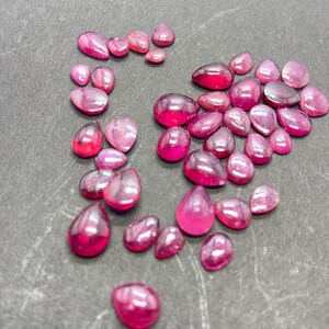 Puede incluir: Una colección de gemas pulidas en forma de lágrima, de varios tamaños. Las gemas son de un rosa intenso y vibrante con un acabado brillante, dispersas sobre una superficie gris oscuro. La disposición sugiere una selección de piedras sueltas, posiblemente para la fabricación de joyas.