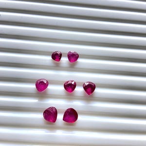 May include: A collection of seven faceted, heart-shaped rubies. The gemstones are a deep, vibrant pinkish-red color and are arranged on a white, ridged surface. The rubies vary slightly in size and shape, showcasing their natural beauty.
