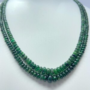 May include: A double-strand necklace featuring small, round, translucent green beads. The beads exhibit varying shades of deep forest green and emerald, with a smooth, polished finish. The strands are arranged in a layered, circular formation.