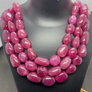 Op de afbeelding: Een ketting met drie strengen met grote, gepolijste, ovaalvormige, roze edelstenen. De ketting wordt getoond op een zwarte mannequin buste. De edelstenen hebben een rijke, dieproze kleur.
