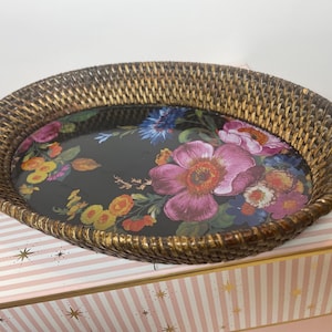 Puede incluir: Una bandeja ovalada con un borde de rat&aacute;n trenzado marr&oacute;n y una base negra plana con un estampado floral colorido. El dise&ntilde;o incluye grandes flores rosas, rojas, amarillas y azules con hojas verdes sobre la superficie oscura de la bandeja.
