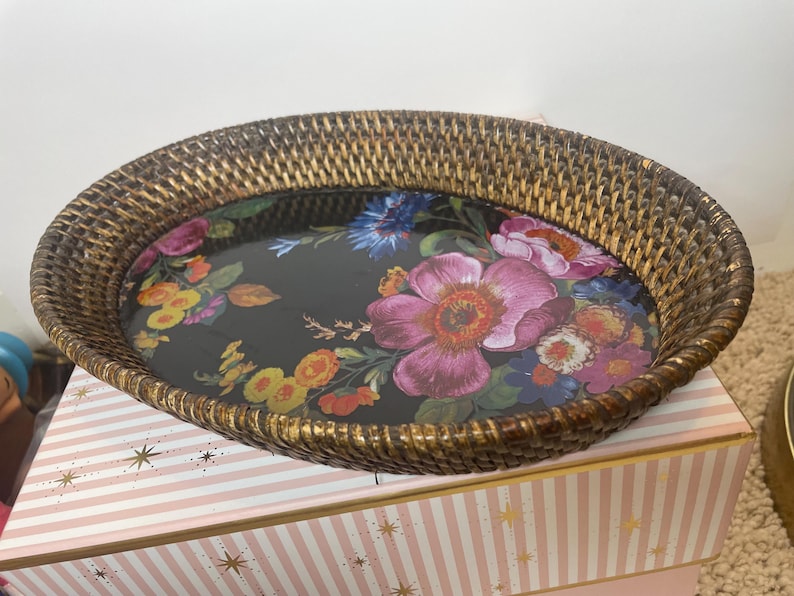 Puede incluir: Una bandeja ovalada con un borde de rat&aacute;n trenzado marr&oacute;n y una base negra plana con un estampado floral colorido. El dise&ntilde;o incluye grandes flores rosas, rojas, amarillas y azules con hojas verdes sobre la superficie oscura de la bandeja.