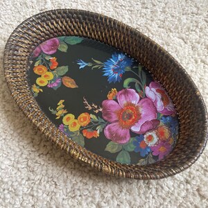 Puede incluir: Una bandeja ovalada con un borde de rat&aacute;n trenzado de color marr&oacute;n oscuro y una base negra con un estampado floral colorido. El dise&ntilde;o incluye grandes flores rosas, naranjas, amarillas y azules con hojas verdes sobre la superficie oscura.